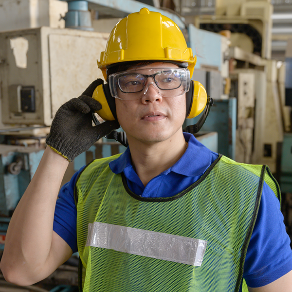 Industrial worker wearing a hard hat, safety goggles, high-visibility vest, and noise-canceling earmuffs in a factory setting.