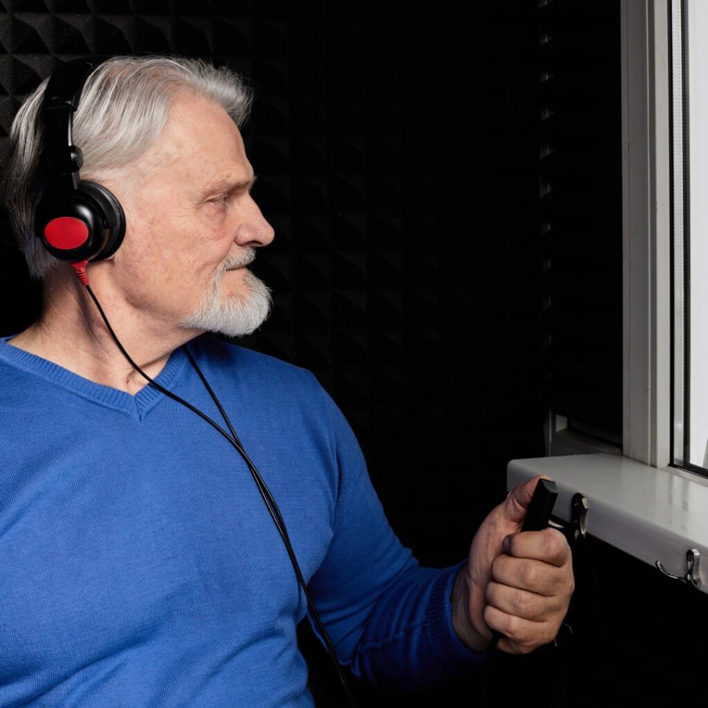 Senior man wearing headphones and holding a response button during an audiometry hearing evaluation inside a sound-treated booth.
