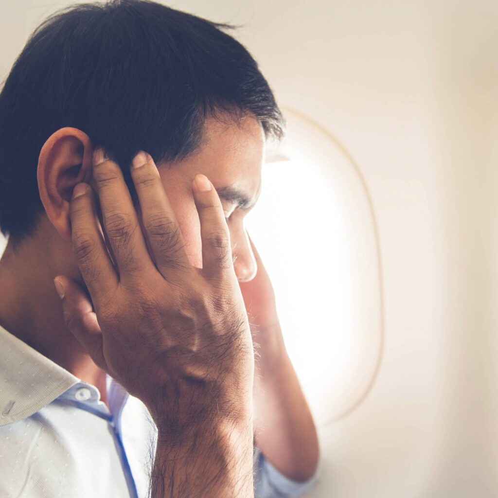 Passenger pressing his hands over his ears inside an airplane cabin, illustrating ear fullness and discomfort from barotrauma during flight.