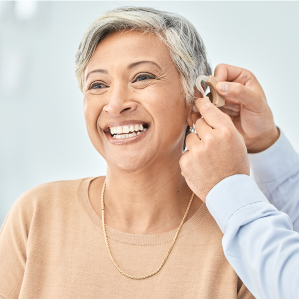 Smiling older woman smiling as a hearing care professional fits a prescription hearing aid behind her ear.