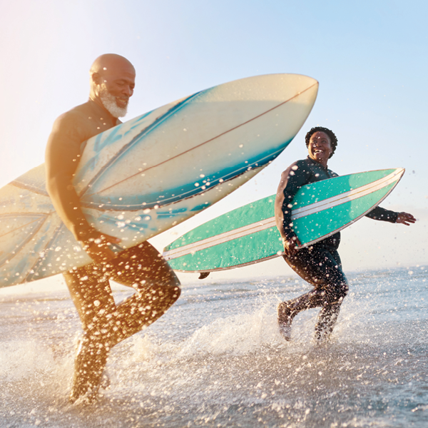 Two adults sprinting through ocean surf with their boards, showing why water‑resistant hearing aids matter for beach and waterside adventures.