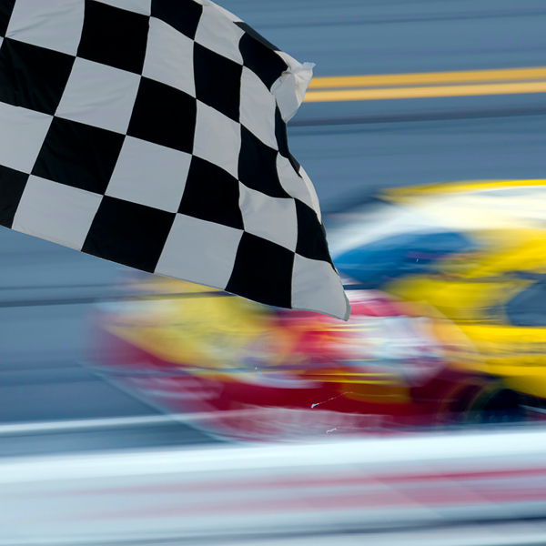 A checkered racing flag waves as a brightly colored stock car speeds past on the NASCAR track, captured with motion blur to convey speed.