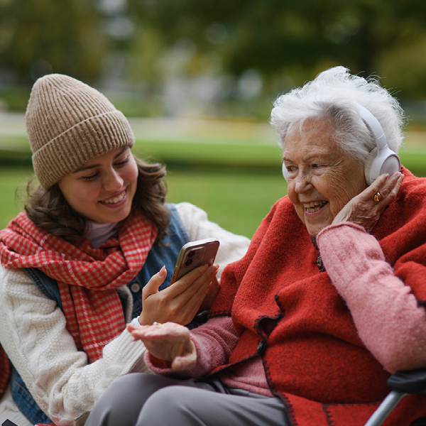 Young woman showing her smartphone to an older woman wearing headphones, both smiling while sitting outside on a cool fall day.