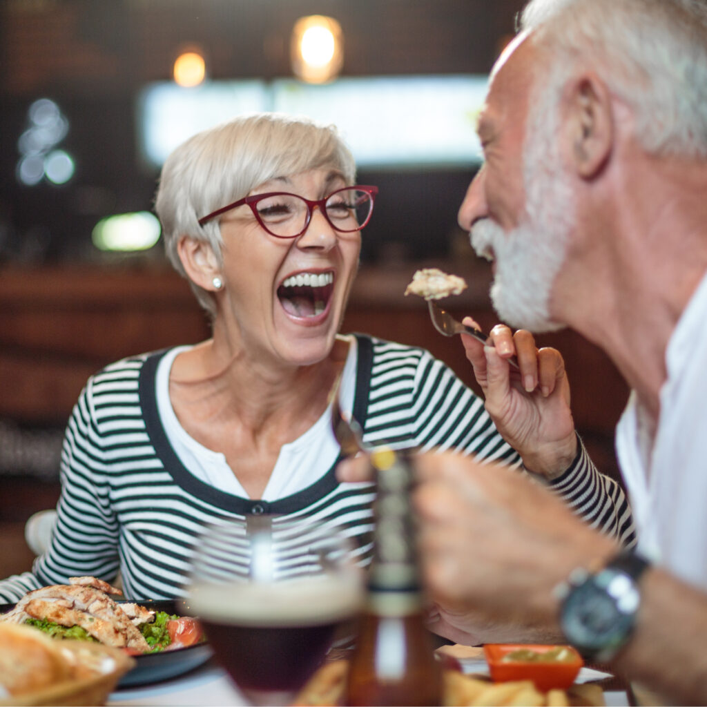 A woman laughing during a meal with a companion at a restaurant, sharing food and conversation.