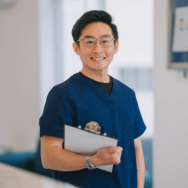 A hearing care professional in navy scrubs stands with a clipboard, smiling warmly inside a clinic.
