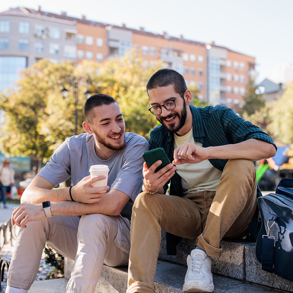 Two young men sit outside together, smiling as they look at a smartphone and enjoy a casual conversation.