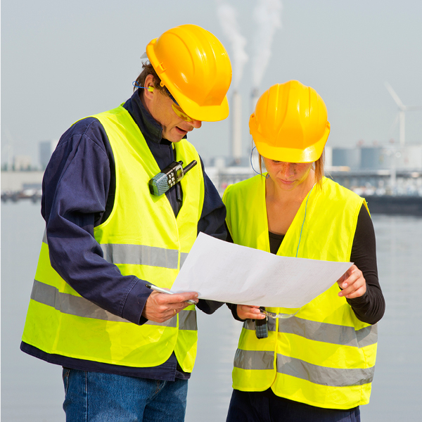 Two construction workers wearing safety vests and hard hats look over a set of plans together outdoors.