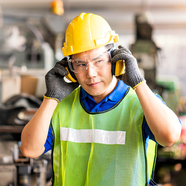 Industrial worker adjusts hearing protection earmuffs in a loud work environment to prevent noise-related hearing loss.