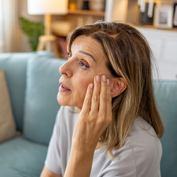 Woman sitting on a couch touching her ear, appearing concerned about ringing or buzzing in her ears.