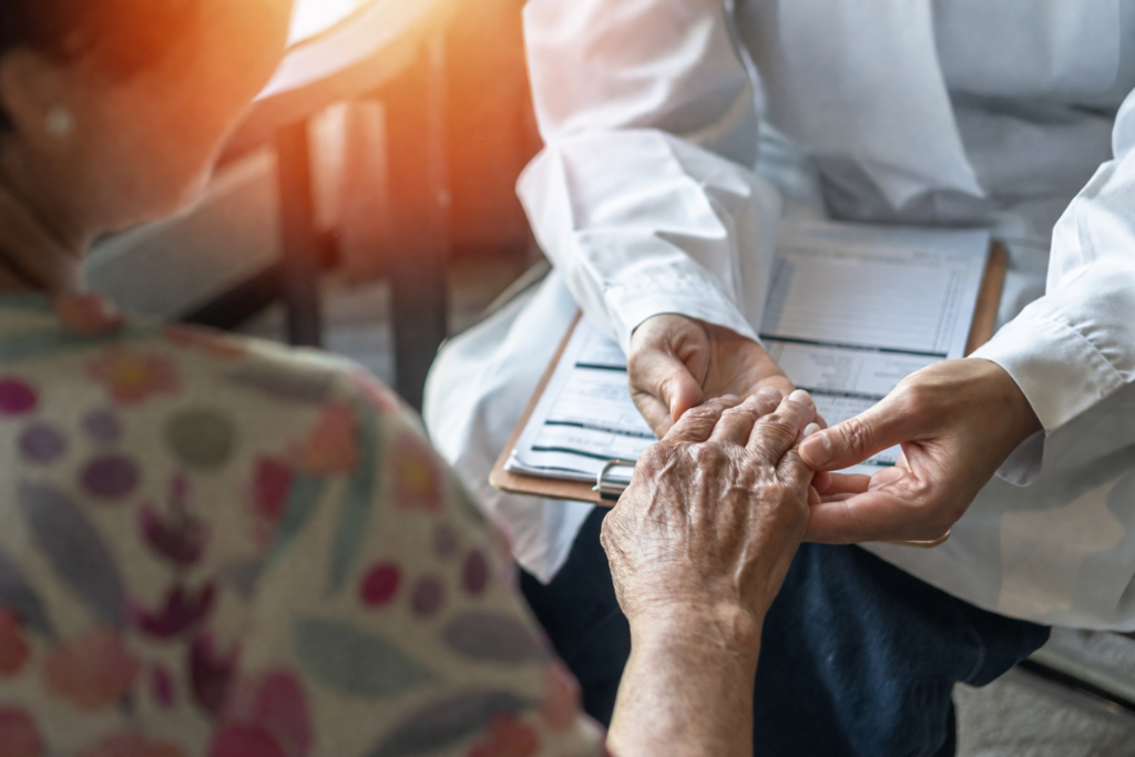 A hearing care professional gently holds the hands of an elderly patient during a medical appointment, with a clipboard visible in the background.