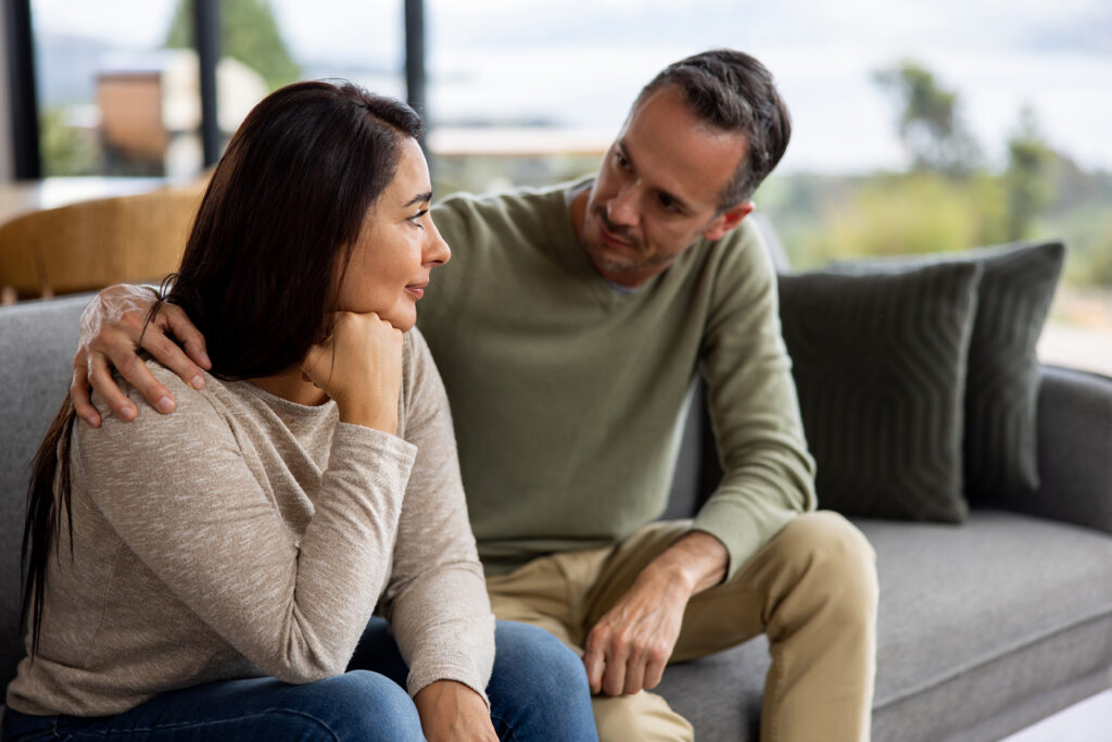 A man places a supportive hand on a woman's shoulder as she sits pensively on a couch, suggesting a caring and empathetic conversation