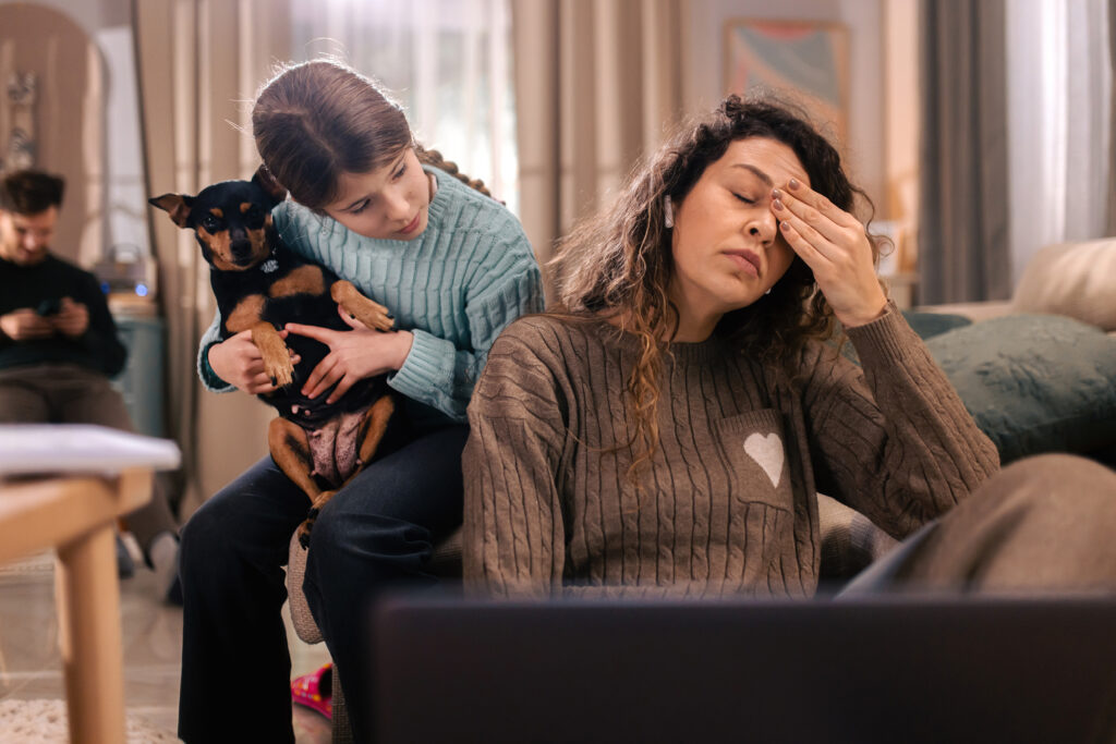 Exhausted woman wearing a hearing aid rubbing her eye while sitting on a couch, with a child holding a dog nearby, illustrating mental fatigue after listening in a busy home environment