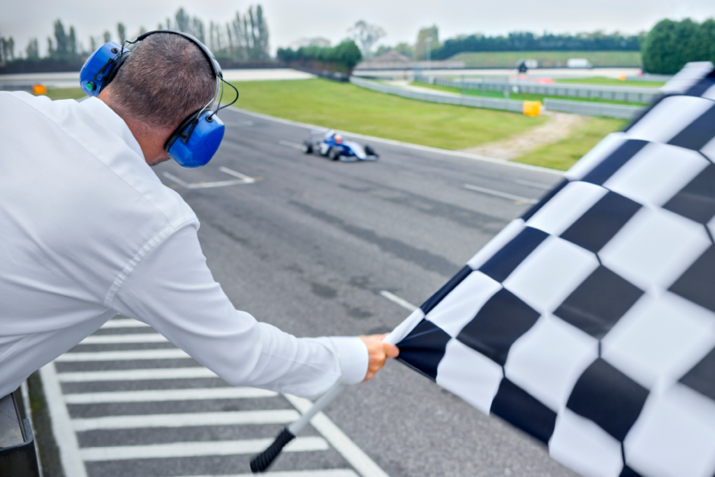 A race official wearing blue ear protection headphones waves a checkered flag as a race car crosses the finish line at an outdoor circuit