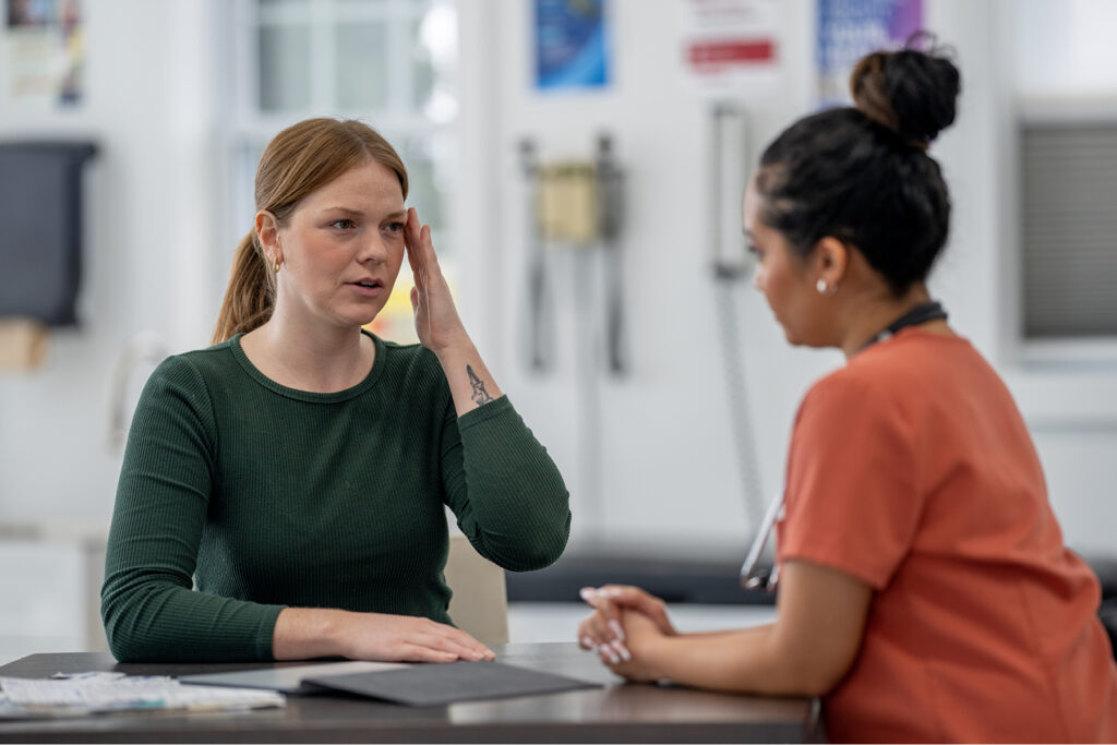 A woman in a green shirt touches her temple while speaking to a hearing care professional wearing orange scrubs.