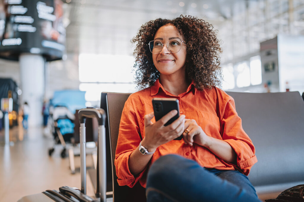 Woman sitting in an airport terminal holding a smartphone and looking up attentively, representing Auracast audio accessibility for travelers.