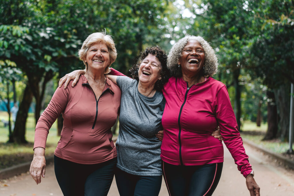 Three smiling older women in workout clothes walking arm in arm and laughing together on a tree lined park path.